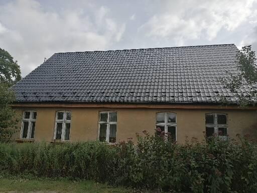 Dom w Człopie z ciemną, odnowioną dachówką i jasną elewacją, częściowo zasłonięty krzewami.