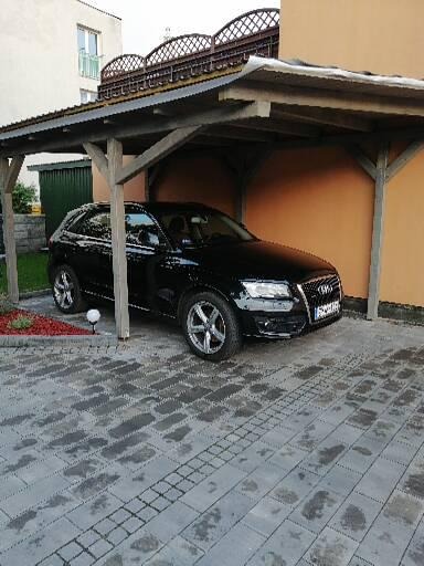 Czarny samochód Audi Q5 parkuje pod drewnianą wiatą garażową z ażurowym wykończeniem dachu, na posesji wyłożonej szarą kostką brukową.