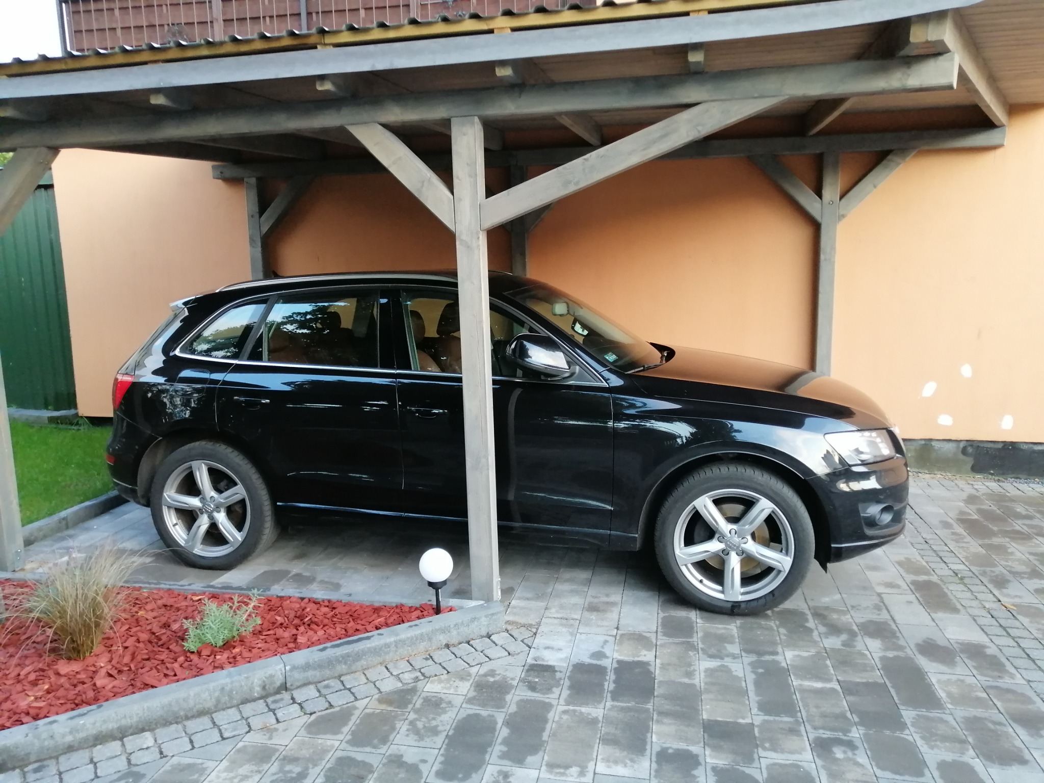 Czarny samochód Audi Q5 zaparkowany pod drewnianą wiatą garażową z oświetleniem punktowym i ozdobnym rabatem z czerwoną korą.