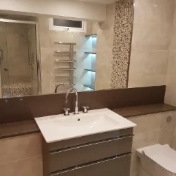 Wykończona łazienka z beżowymi płytkami, podwieszaną szafką z umywalką, dużym lustrem i kabiną prysznicową w tle, z widocznymi mozaikami.