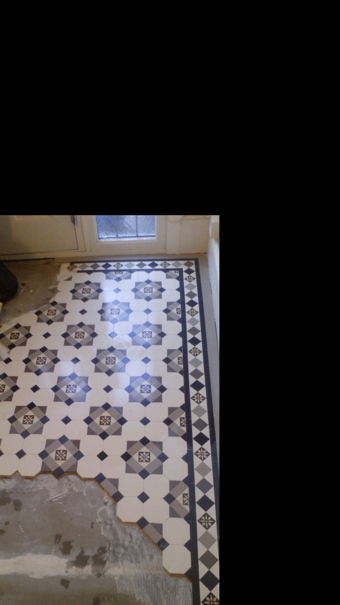 Fragment podłogi w trakcie układania płytek w stylu retro, widoczne niedokończone krawędzie i wzór z motywem kwiatowym w odcieniach szarości i bieli.