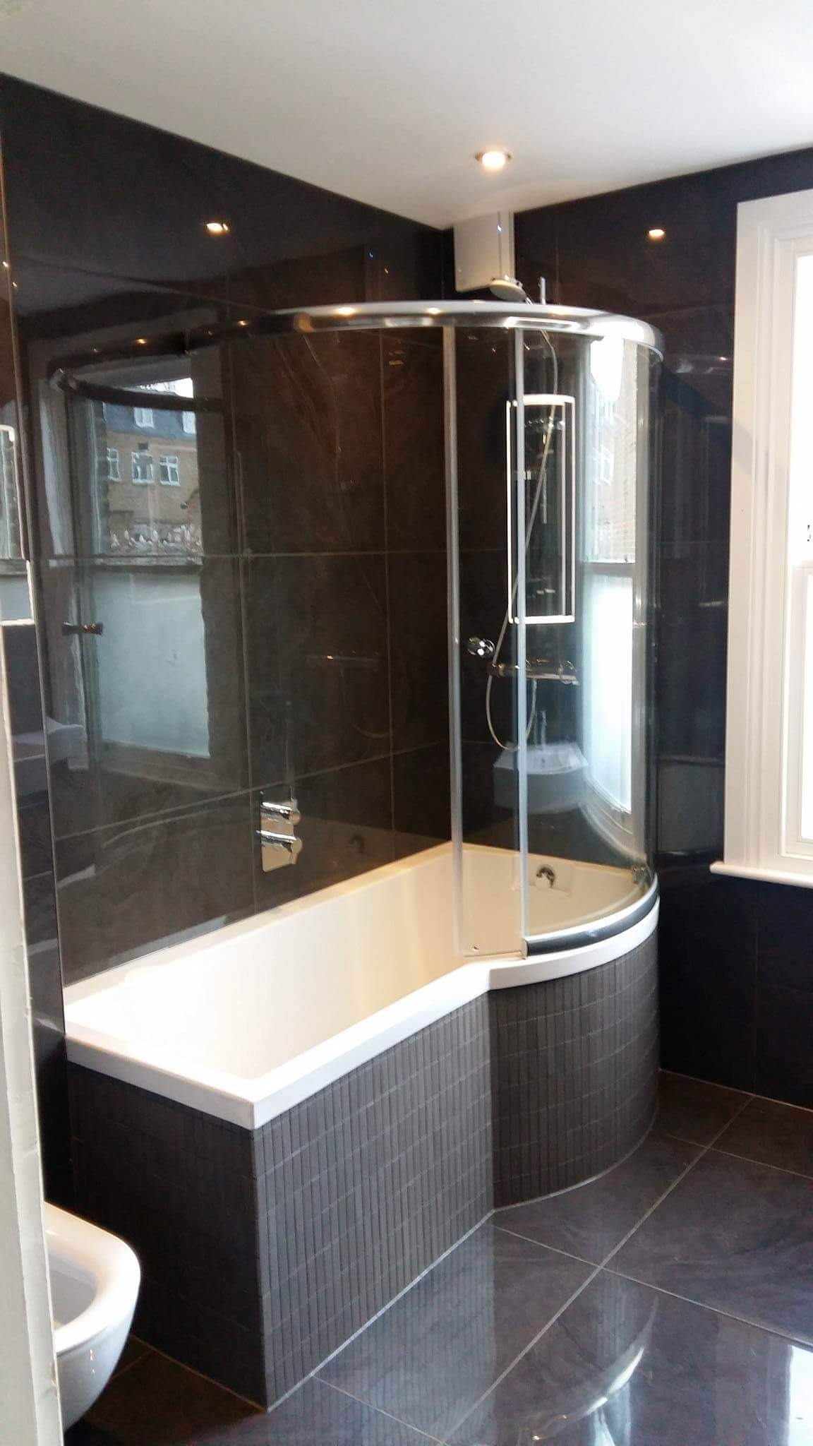 Nowoczesna łazienka z wanną obudowaną mozaiką w odcieniach szarości, przeszklona kabina prysznicowa z chromowanymi detalami, ciemne płytki na ścianach i podłodze, okno z białymi ramami.