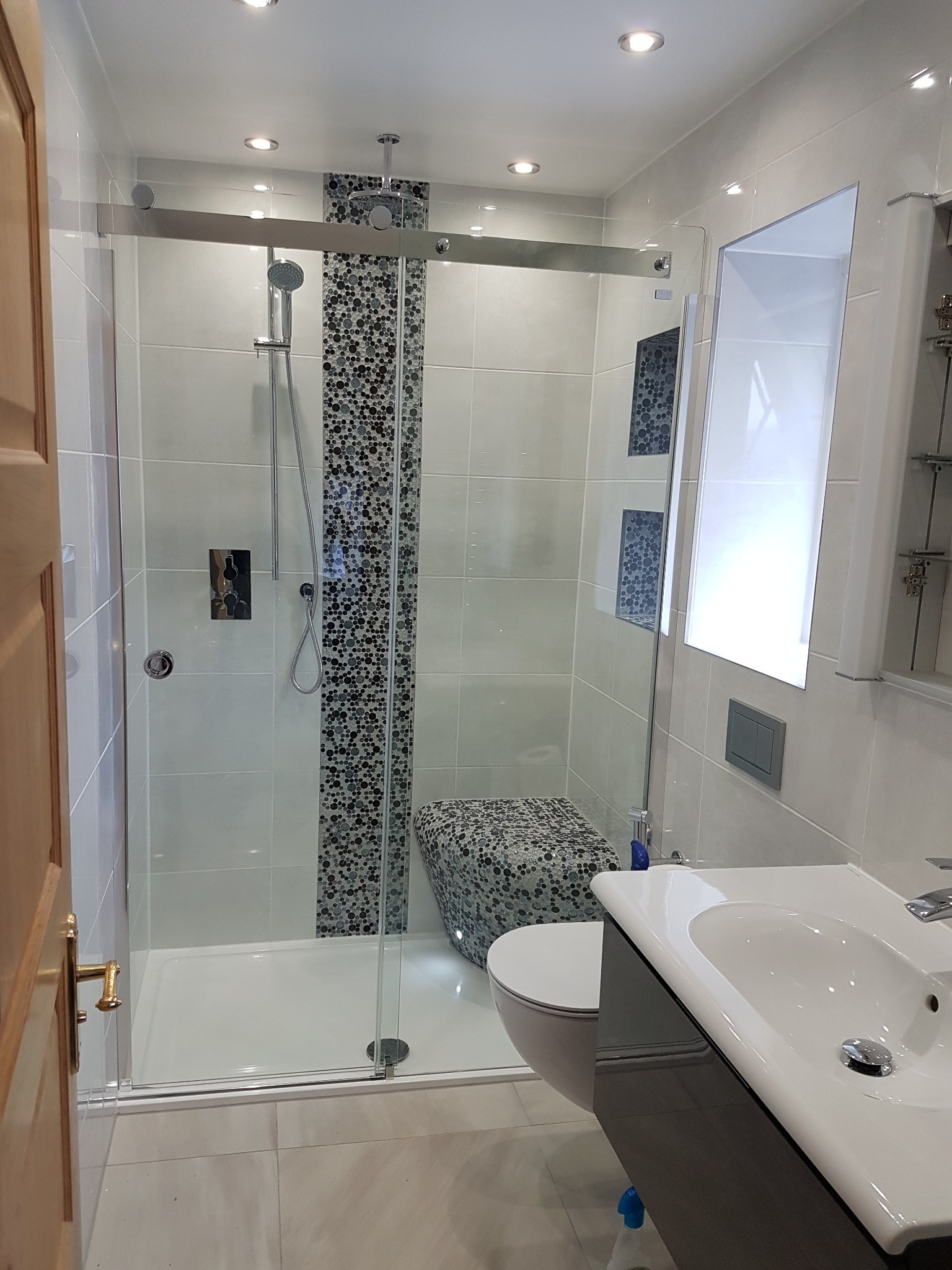 Nowoczesna łazienka z kabiną prysznicową typu walk-in, wykończona jasnymi płytkami i mozaiką na ścianie, widoczna umywalka z szafką i toaleta z mozaikowym siedziskiem, oświetlenie punktowe w suficie.