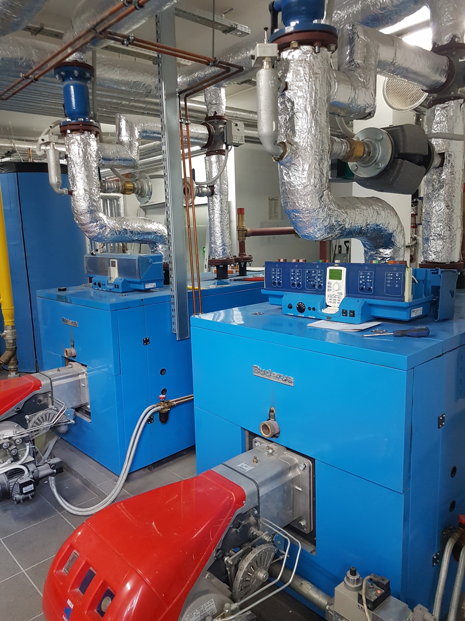 Dwa niebieskie kotły grzewcze firmy Buderus z czerwonymi palnikami Riello, połączone rurami z izolacją aluminiową, z panelem sterowania na górze, na szarej podłodze.