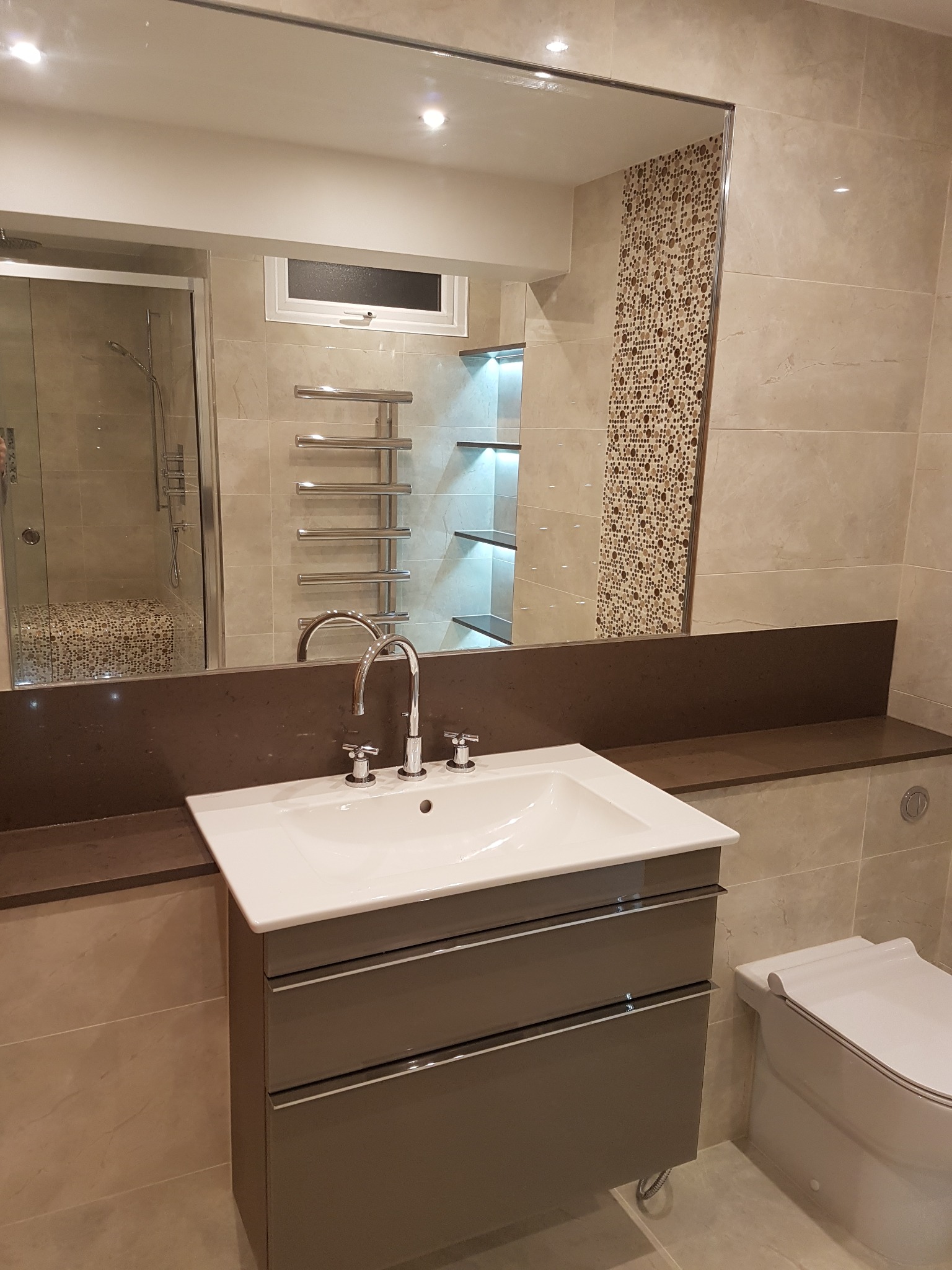 Wykończona łazienka z beżowymi płytkami, podwieszaną szafką z umywalką, dużym lustrem i kabiną prysznicową w tle, z widocznymi mozaikami.