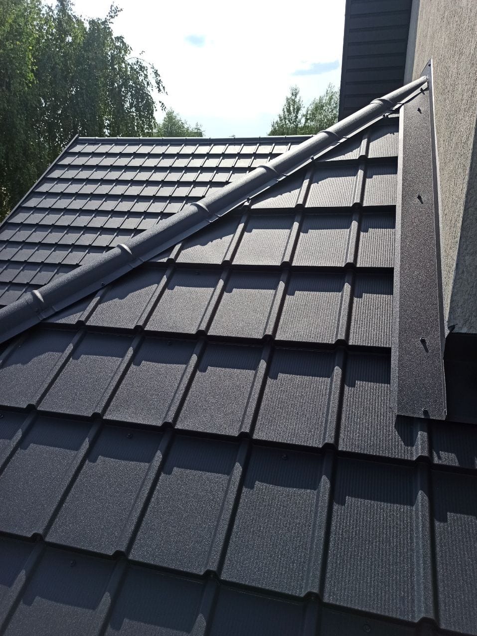 Nowy, grafitowy dach z blachodachówki modułowej, z widocznymi obróbkami blacharskimi przy ścianie budynku, w słoneczny dzień.