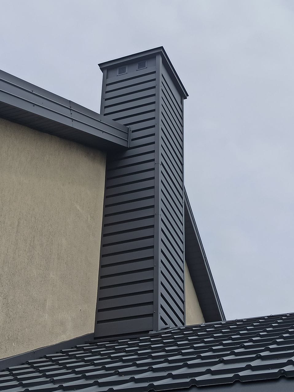 Szary komin z blachy trapezowej z poziomymi szczelinami wentylacyjnymi na dachu pokrytym szarą blachodachówką, widok z dołu.