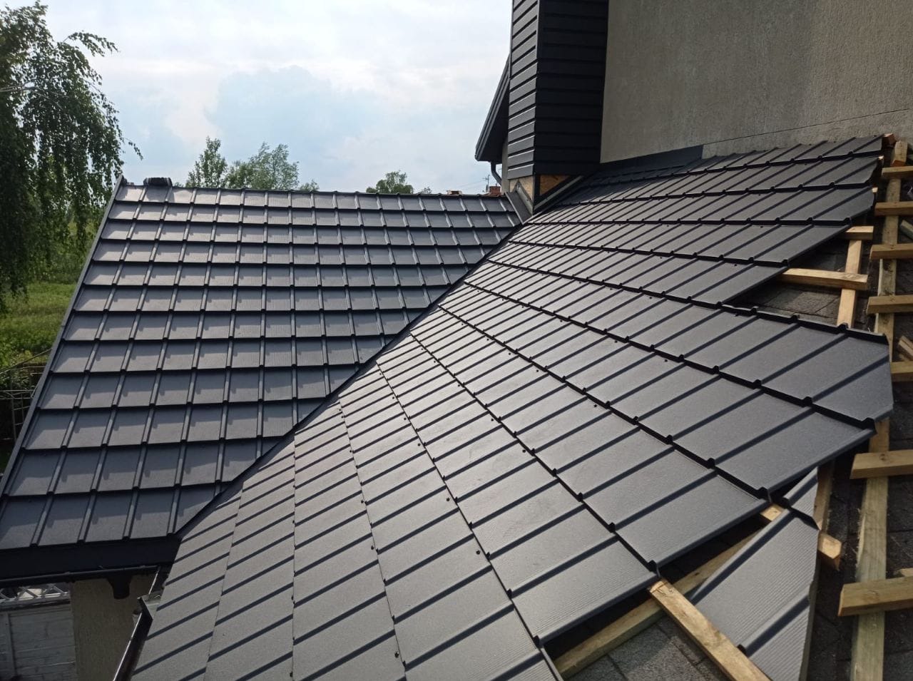 Fragment dachu z czarnej dachówki modułowej, widoczne elementy konstrukcji więźby dachowej po prawej stronie oraz komin w tle.