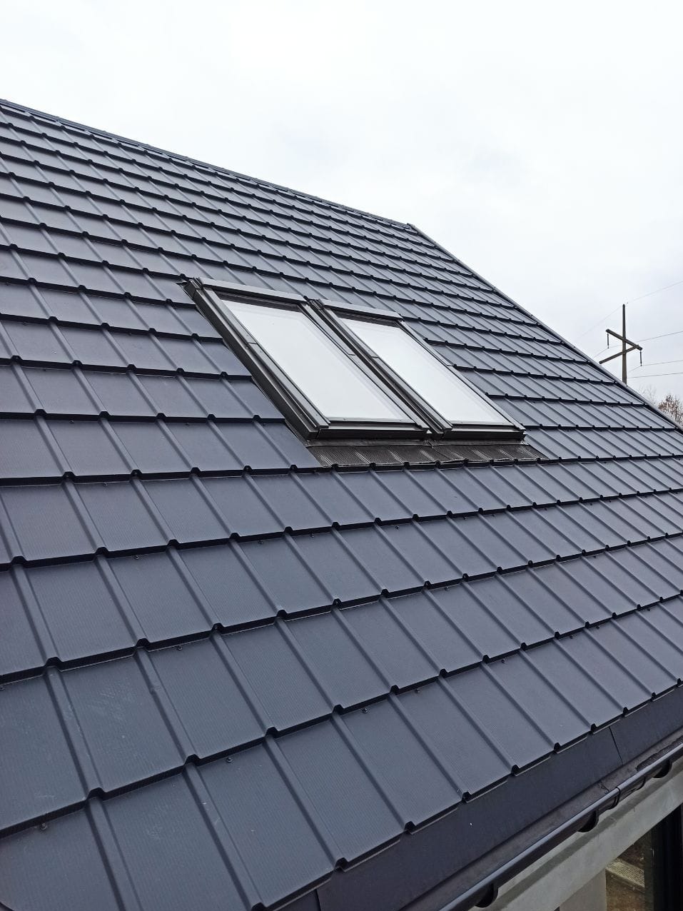 Szary dach z blachy trapezowej z dwoma oknami dachowymi, widoczne połączenia blach i mocowania.