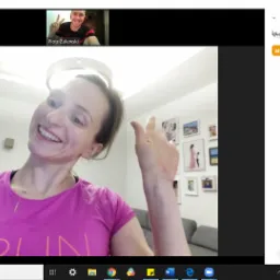 Kobieta w r&oacute;żowej koszulce z napisem 'RUN' uśmiecha się i macha ręką podczas wideokonferencji, w tle ściana z ramkami zdjęć, sesja online z trenerem personalnym w Warszawie.