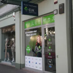 Fasada punktu partnerskiego CUK Ubezpieczenia z ekspozycją reklamową w oknach, prezentująca szeroki zakres ubezpieczeń: auto, dom, życie, podróże, firmy, floty, rowery i NNW.