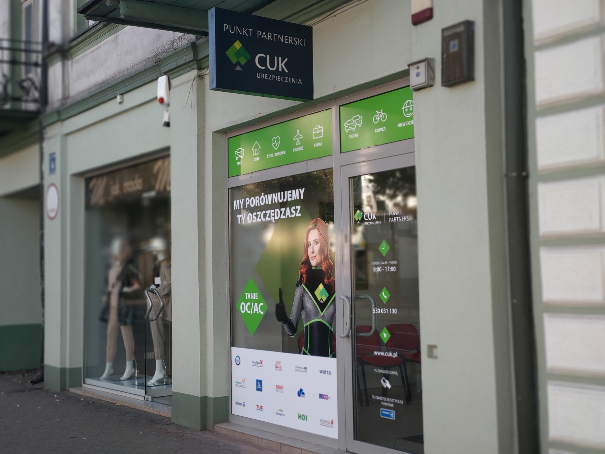 Fasada punktu partnerskiego CUK Ubezpieczenia z ekspozycją reklamową w oknach, prezentująca szeroki zakres ubezpieczeń: auto, dom, życie, podróże, firmy, floty, rowery i NNW.
