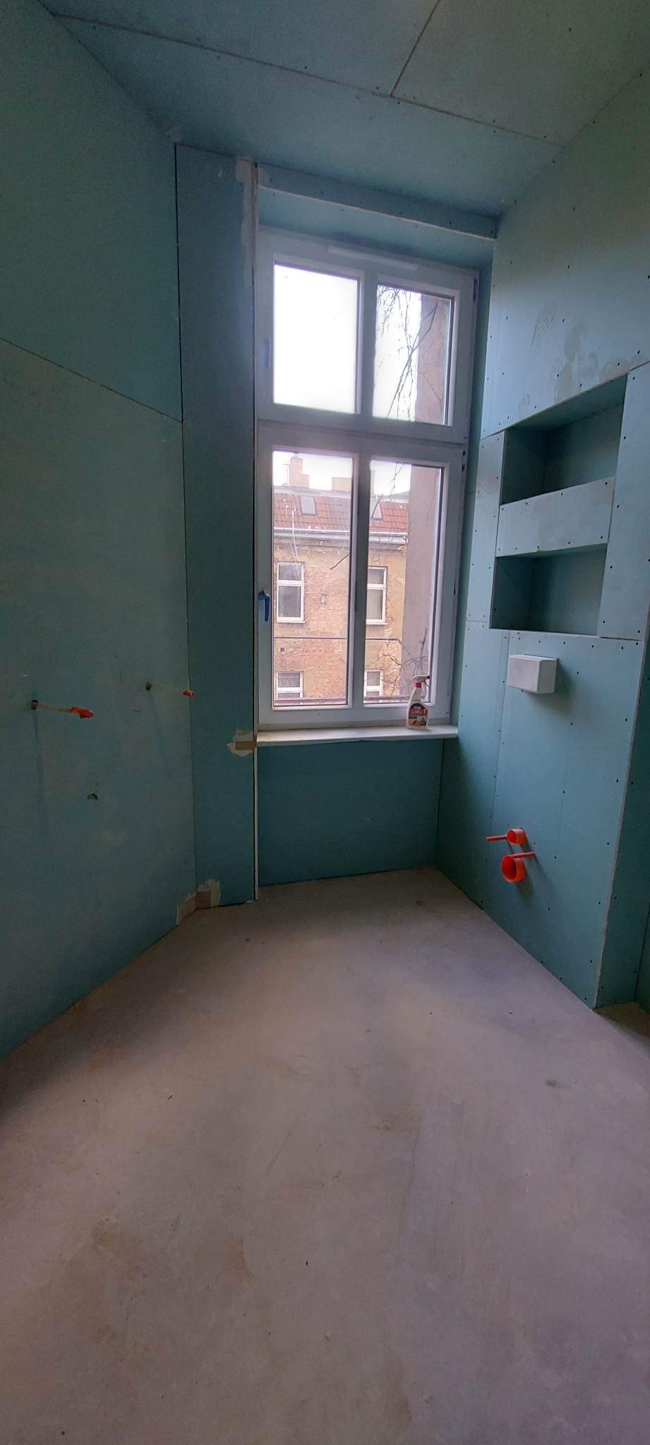 Łazienka w trakcie remontu: ściany obłożone płytami kartonowo-gipsowymi, widoczne instalacje hydrauliczne i wnęki, okno z widokiem na budynek mieszkalny.
