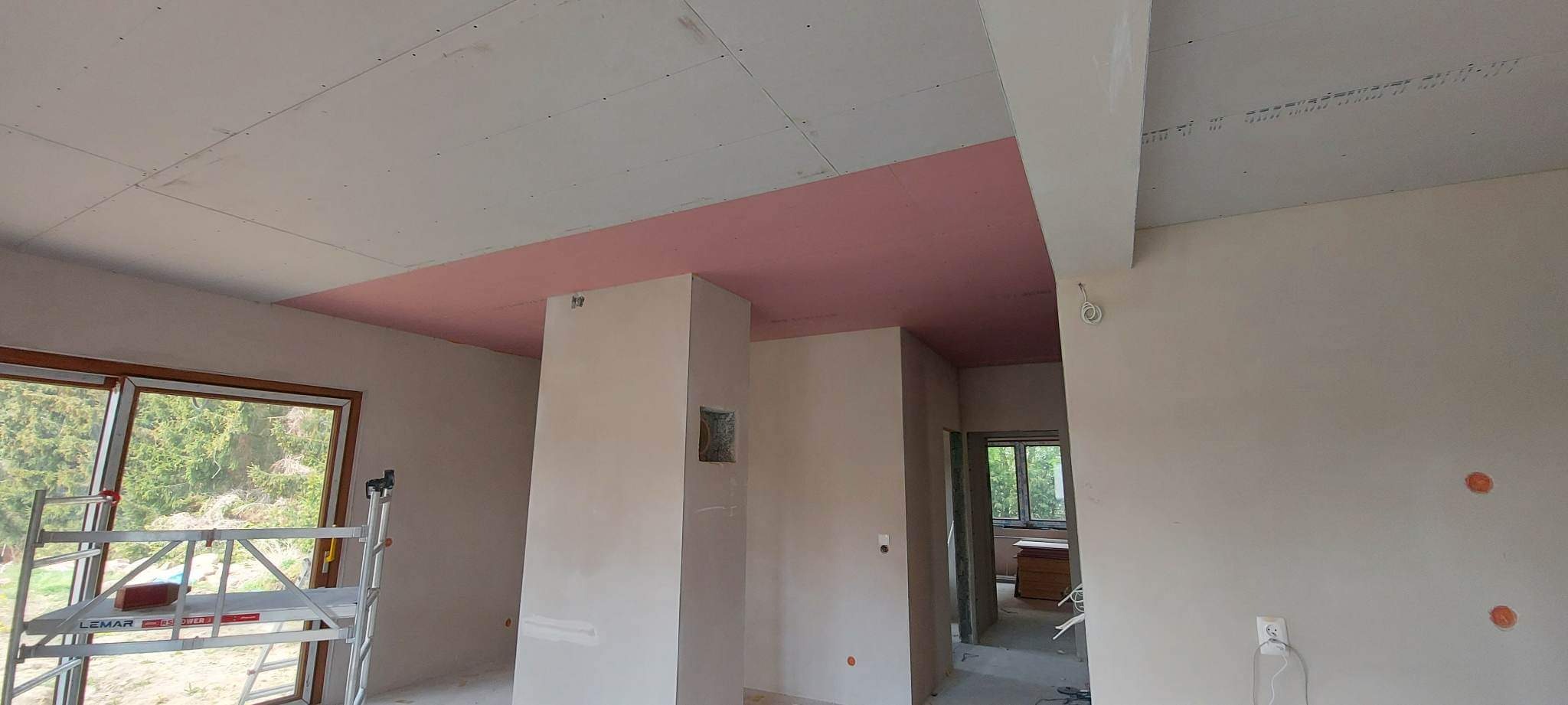 Wnętrze w trakcie prac wykończeniowych: sufit częściowo pokryty czerwoną płytą, ściany przygotowane do malowania, widoczny fragment rusztowania przy oknie.