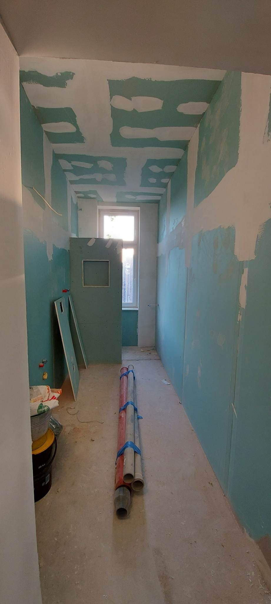 Wąskie pomieszczenie w trakcie prac wykończeniowych: ściany i sufit pokryte częściowo płytami gipsowo-kartonowymi, widoczne szpachlowanie łączeń, na podłodze leżą rury PCV związane niebieską taśmą,...