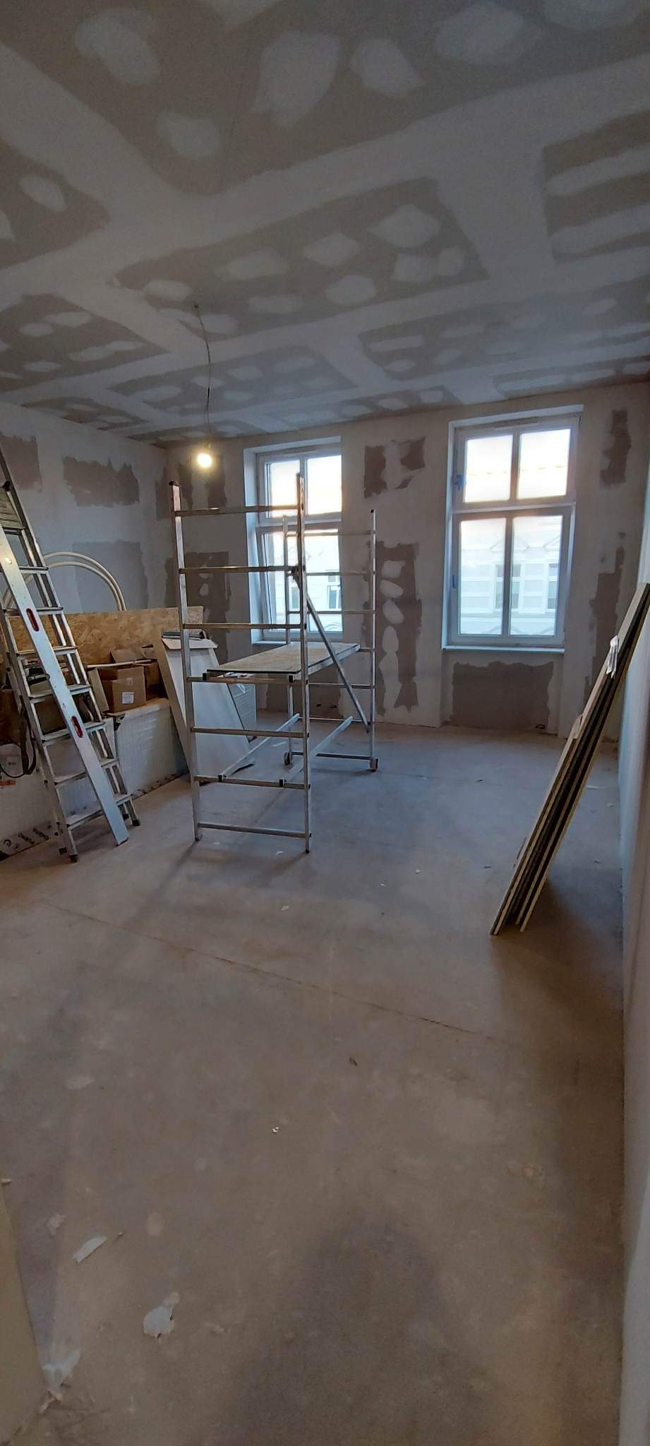 Pokój w trakcie prac wykończeniowych, widoczne rusztowania, płyty i drabina, ściany przygotowane do malowania, sufit z zamontowaną płytą gipsowo-kartonową przed szpachlowaniem.