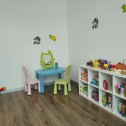 Umeblowany pokój dziecięcy z niskim stolikiem, krzesełkami, regałem pełnym zabawek i dekoracjami ściennymi z motywem żab i żółwi.