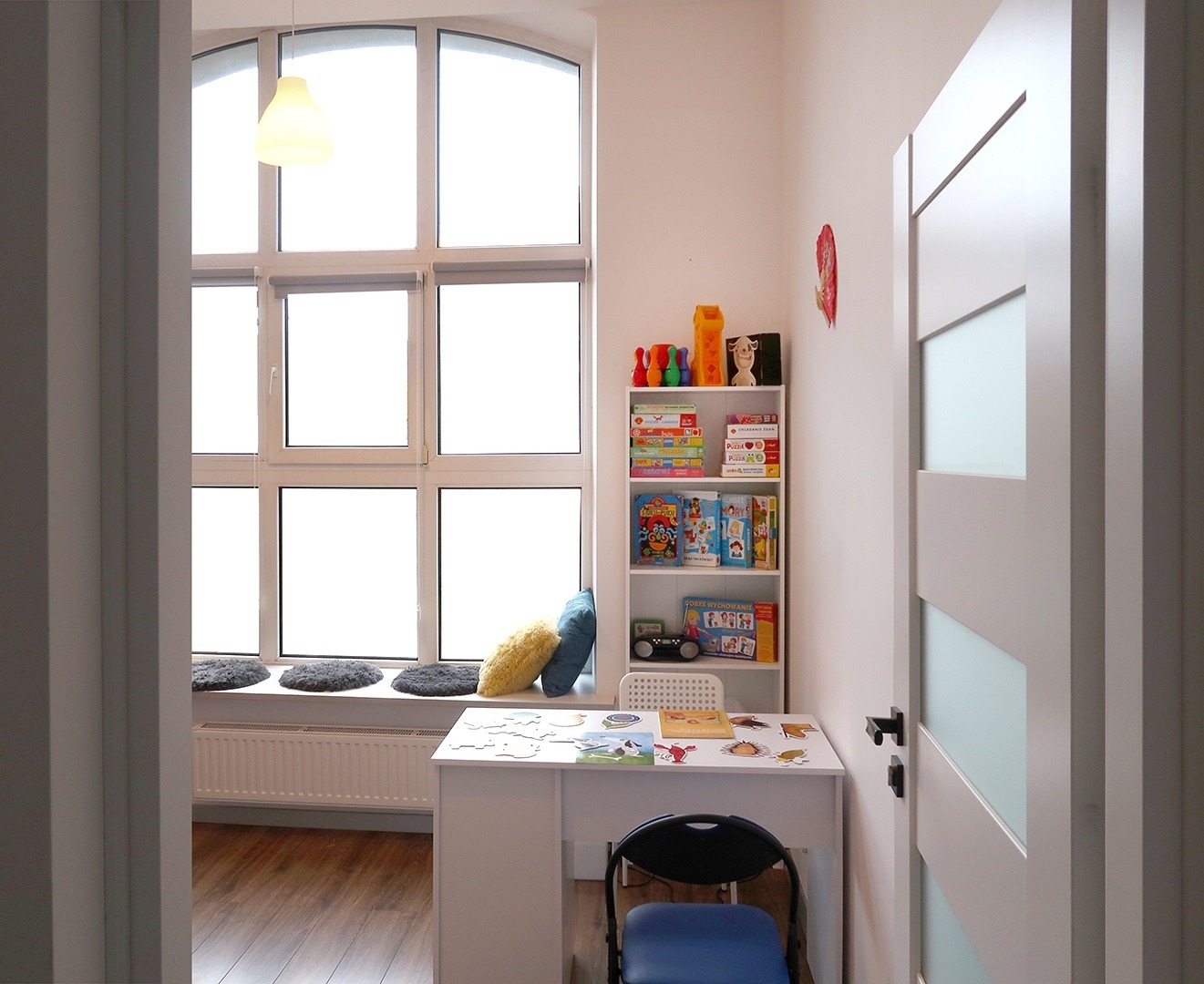 Jasny gabinet z dużym oknem, biurkiem, krzesłem i regałem z zabawkami edukacyjnymi, przygotowany do zajęć terapeutycznych.