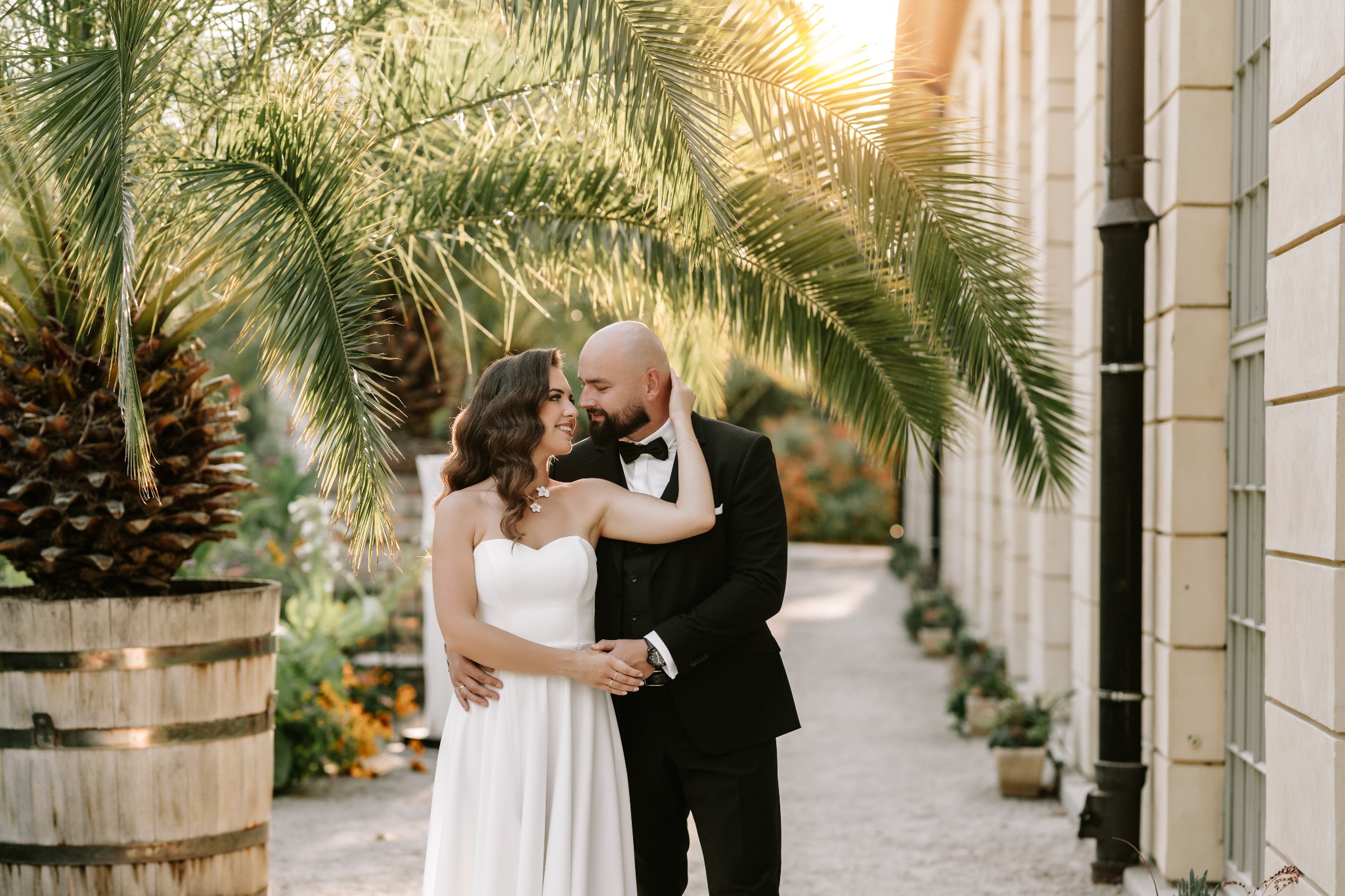 Para młoda w objęciach pod palmami w słoneczny dzień. Panna młoda w białej sukni, pan młody w czarnym smokingu. Romantyczna sceneria ślubna z architekturą w tle.