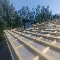 Konstrukcja dachu z drewnianymi łatami i kominem w trakcie budowy, widok z dołu na tle drzew i błękitnego nieba.