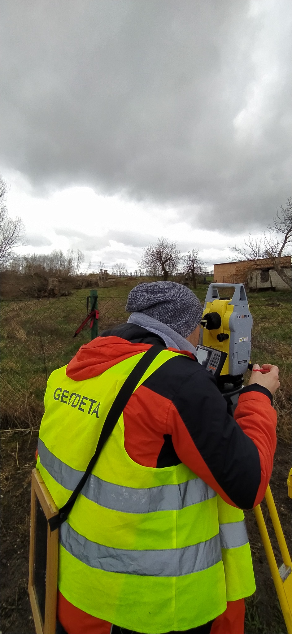 Osoba w odblaskowej kamizelce z napisem GEODETA, obsługująca żółty teodolit na trójnogu, na tle pochmurnego nieba i terenu z drzewami.