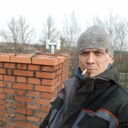 Murarz z papierosem w ustach na tle komina z cegły i metalowego wylotu spalin