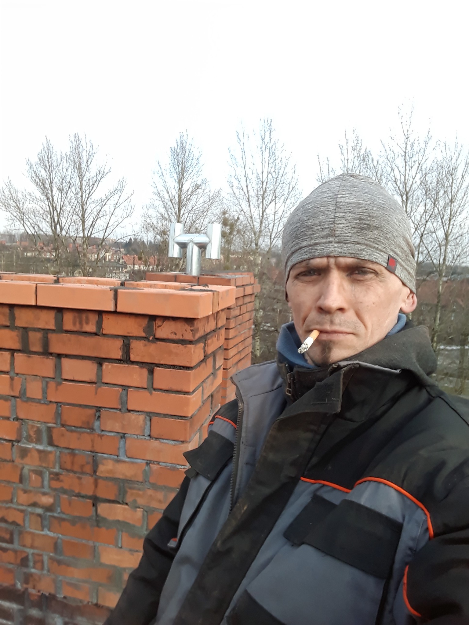 Murarz z papierosem w ustach na tle komina z cegły i metalowego wylotu spalin