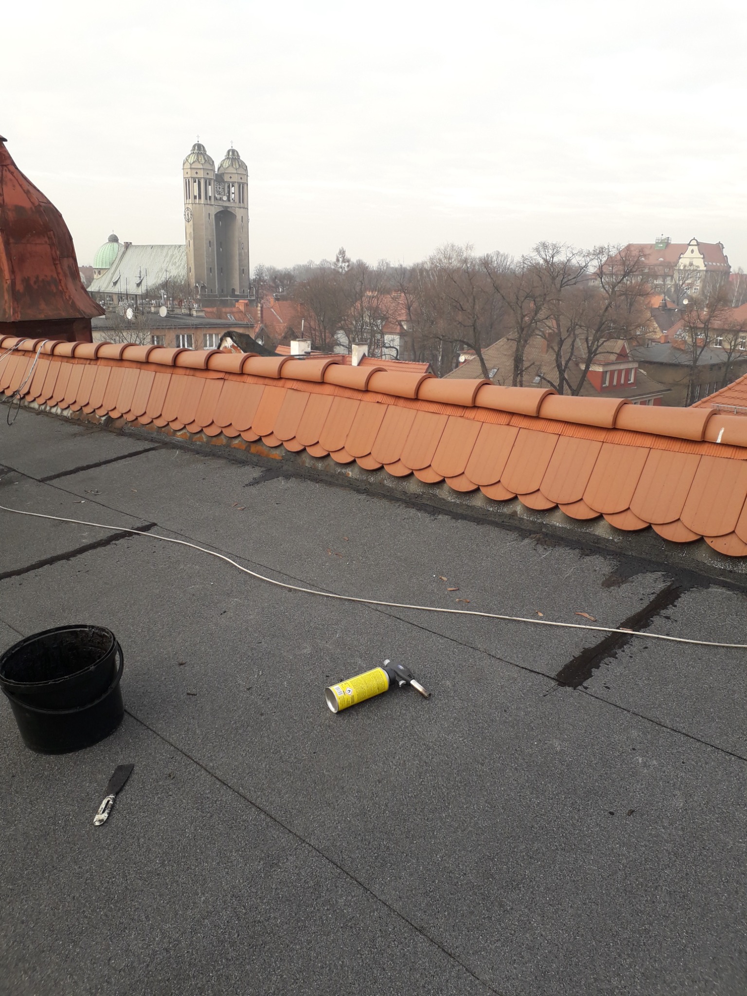 Papa termozgrzewalna na dachu z widocznym palnikiem, wiadrem i szpachelką, w tle zabudowa miejska i kościół.