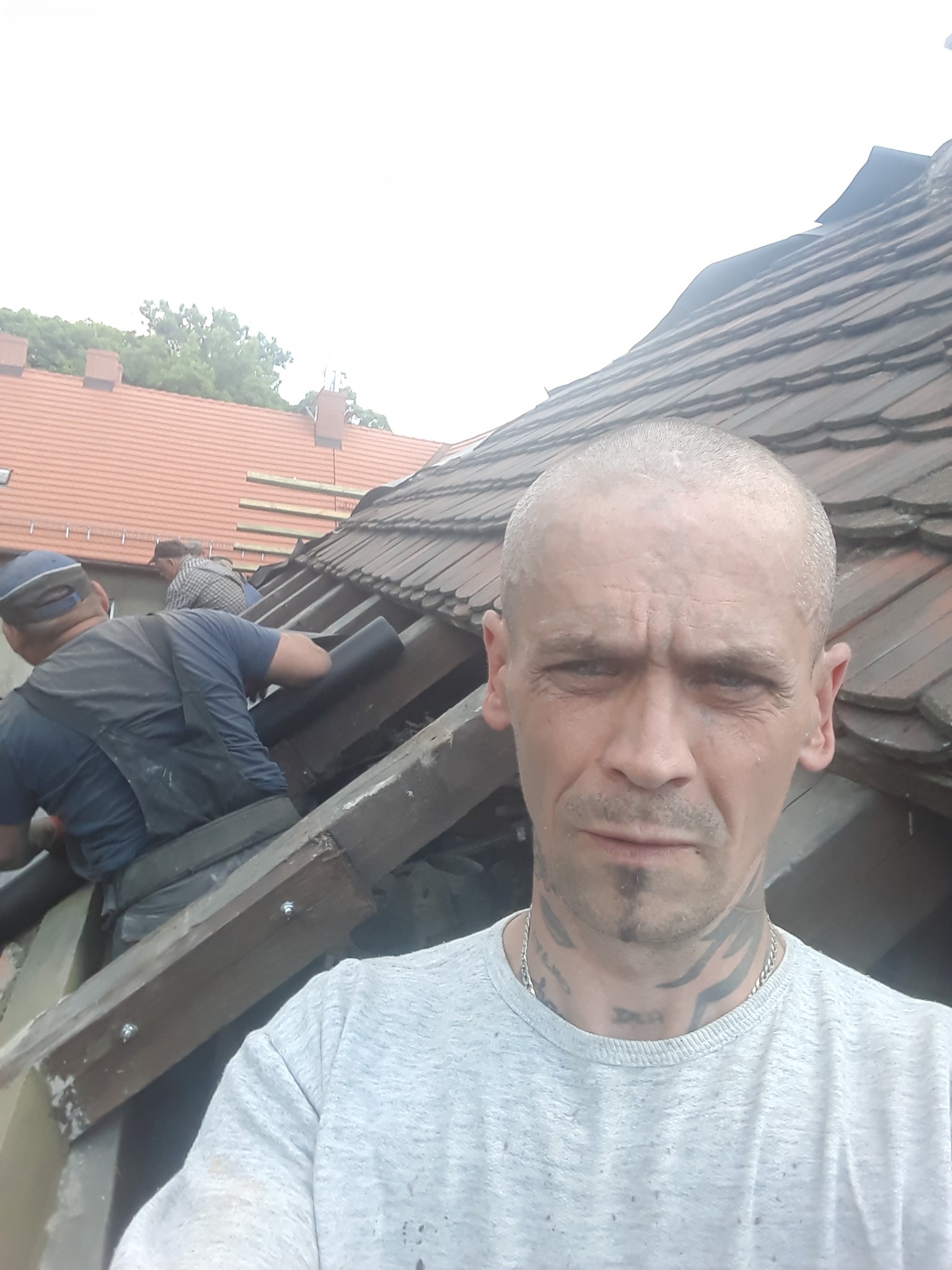 Mężczyzna z tatuażami na szyi robiący selfie na tle remontowanego dachu z widocznymi pracownikami i elementami konstrukcyjnymi