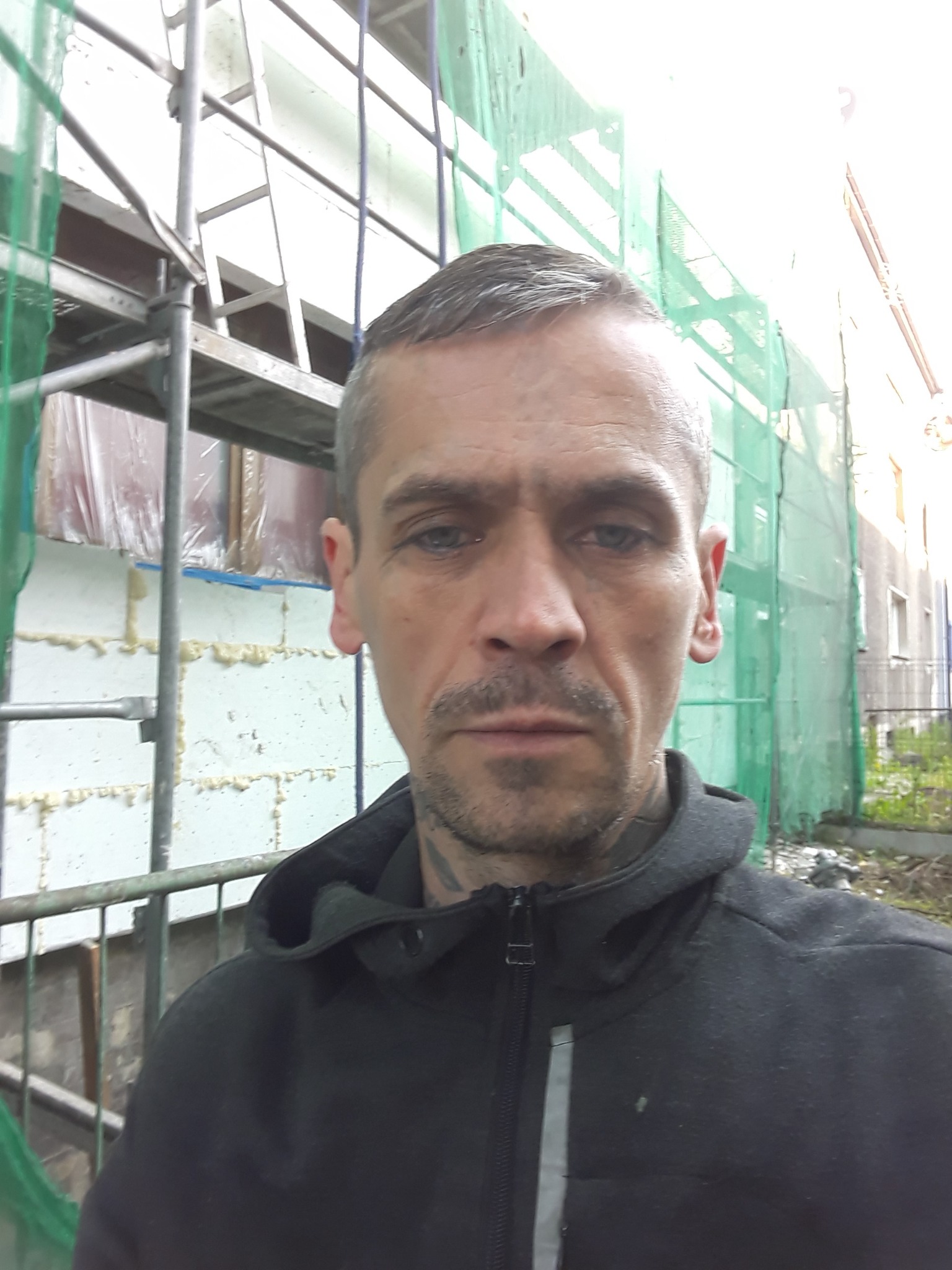 Mężczyzna w bluzie na tle rusztowania i ocieplonej ściany budynku, z widoczną siatką ochronną.