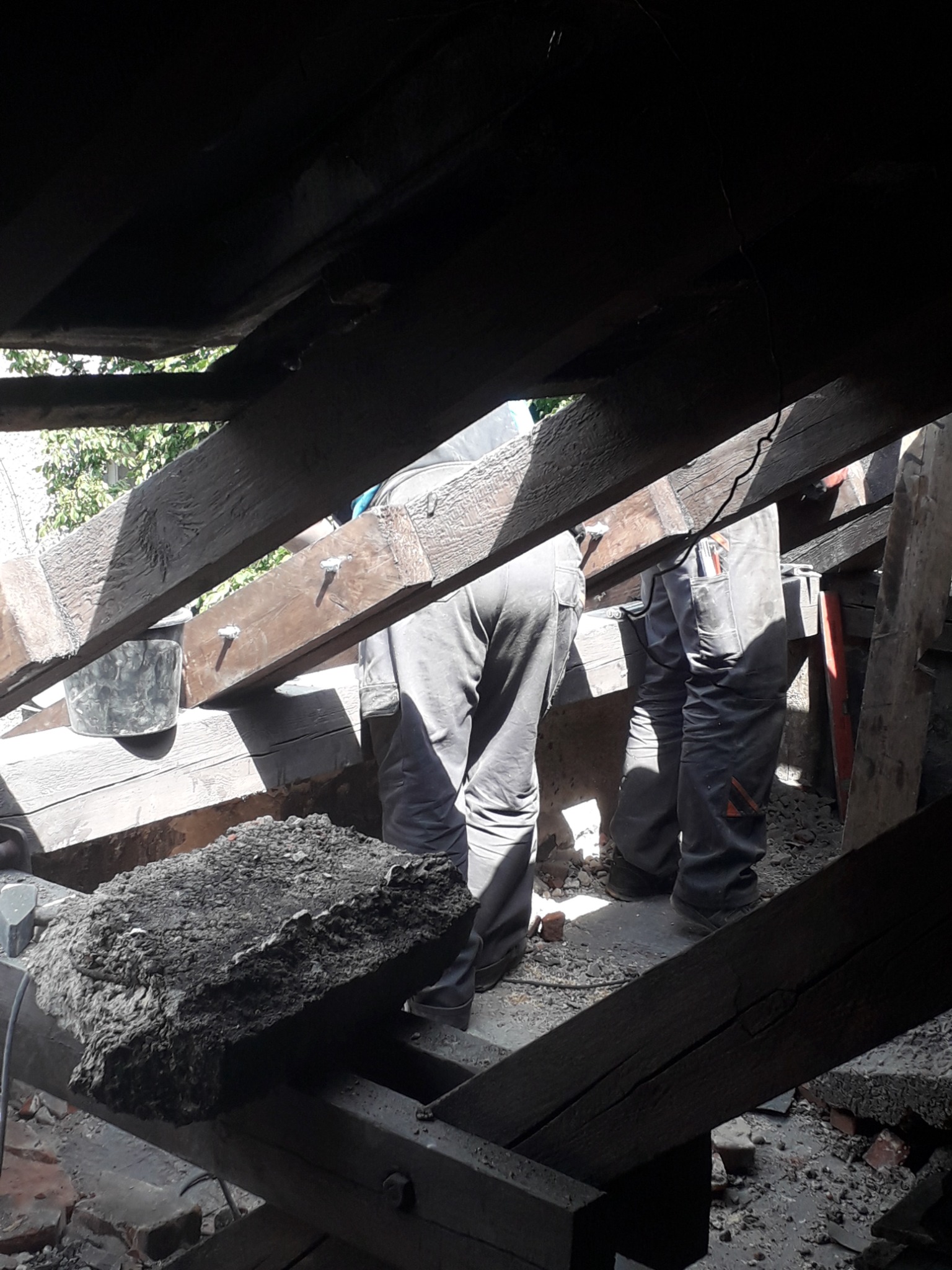 Ekipa remontowa w trakcie prac na poddaszu, widoczne drewniane belki konstrukcyjne, gruz i narzędzia.