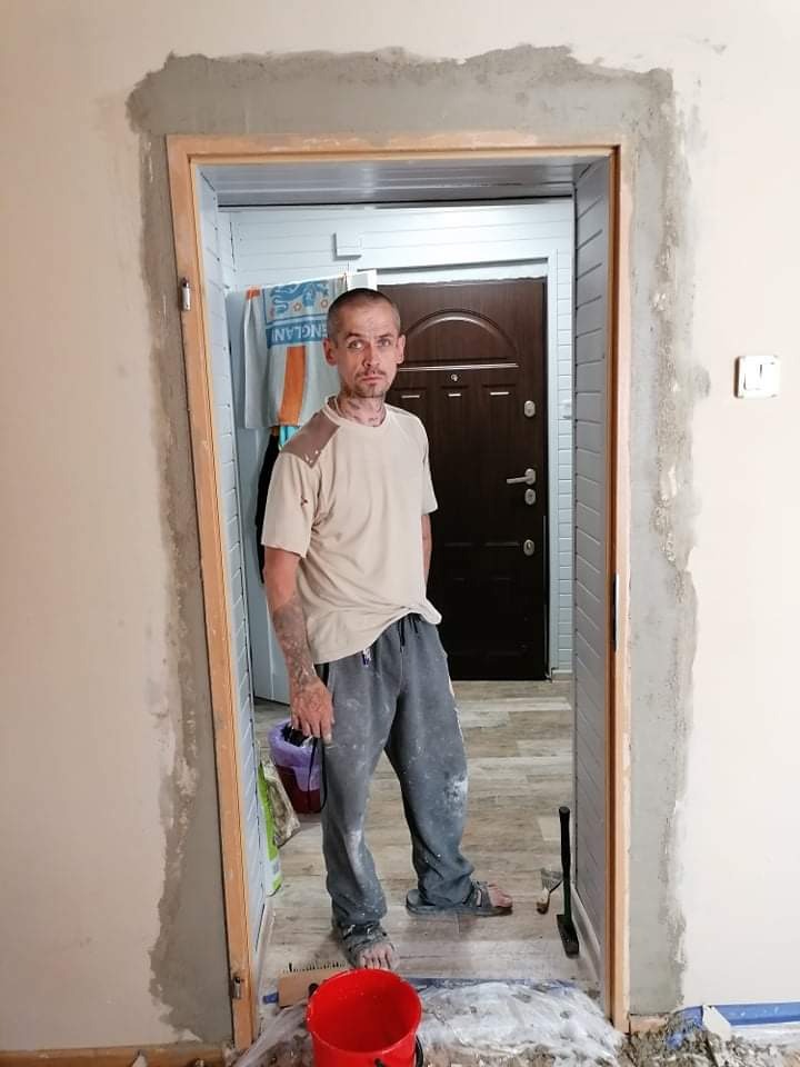 Mężczyzna w ubrudzonych farbą spodniach stoi w przejściu z ościeżnicą po szpachlowaniu, w ręku trzyma pojemnik z farbą, widoczne narzędzia i wiadro.