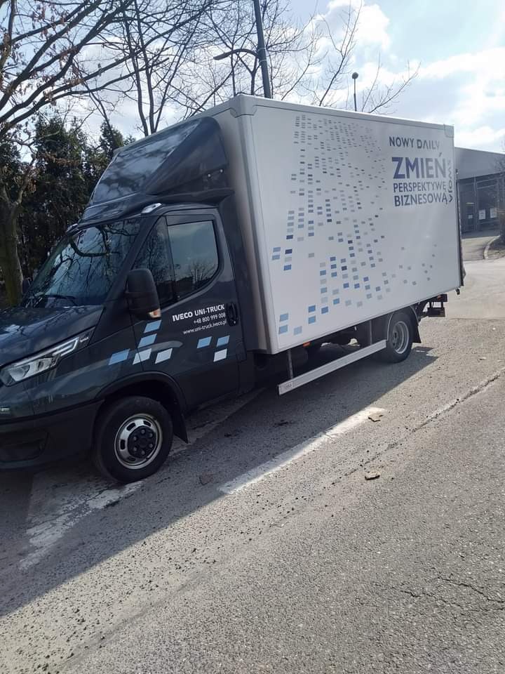Szary samochód dostawczy Iveco Uni-Truck z reklamą 'Nowy Daily - Zmień perspektywę biznesową' na białej zabudowie, zaparkowany na poboczu drogi w słoneczny dzień.