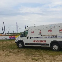 Biały samochód dostawczy z reklamą usług motoryzacyjnych Auto-Orunia na tle toru wyścigowego, z widocznymi banerami reklamowymi i flagami.