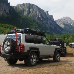 Srebrny samochód terenowy z bagażnikiem dachowym i kołem zapasowym na tle górskiego krajobrazu. Na dachu namiot. Widoczny podnośnik typu hi-lift.