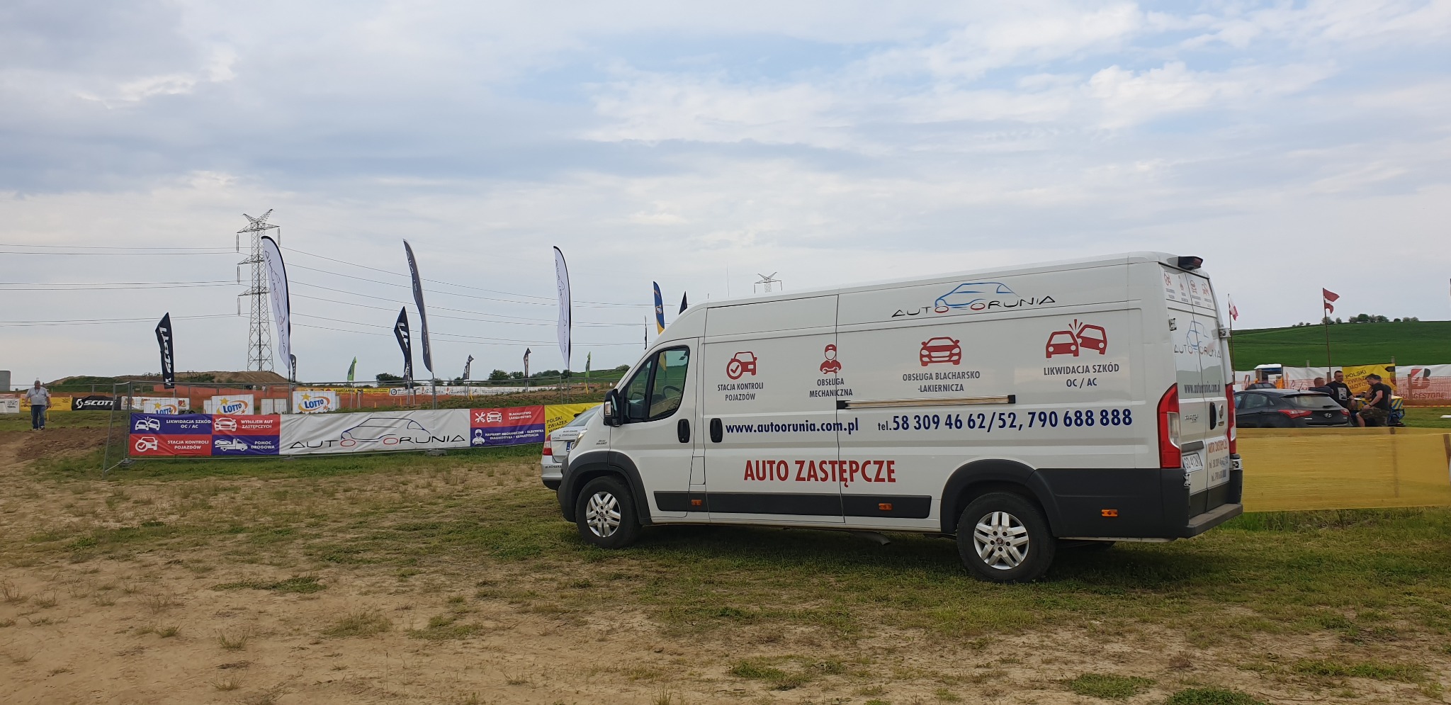 Biały samochód dostawczy z reklamą usług motoryzacyjnych Auto-Orunia na tle toru wyścigowego, z widocznymi banerami reklamowymi i flagami.