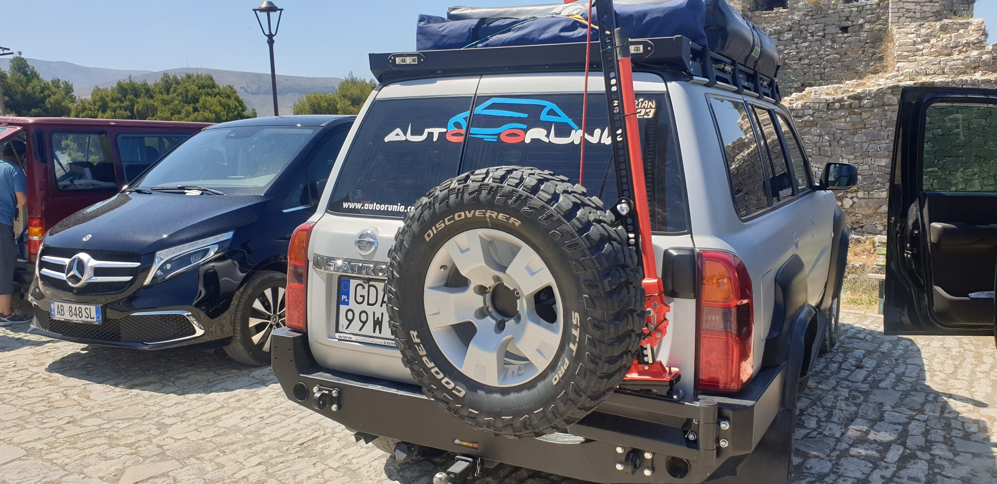 Szary Nissan Patrol z bagażnikiem dachowym, kołem zapasowym zamontowanym z tyłu oraz podnośnikiem Hi-Lift, zaparkowany na brukowanej nawierzchni, widoczny logotyp 'AUTORUNIA' na tylnej szybie.