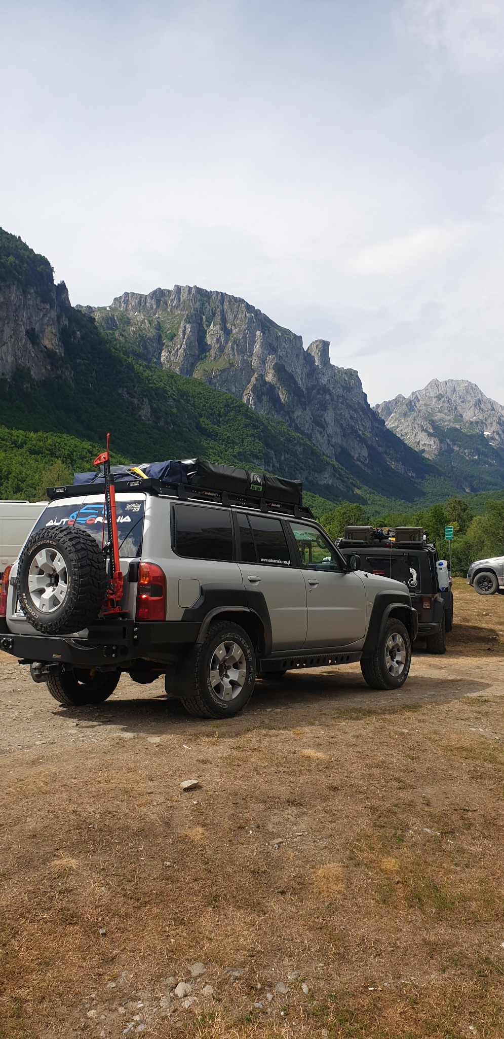 Srebrny samochód terenowy z bagażnikiem dachowym i kołem zapasowym na tle górskiego krajobrazu. Na dachu namiot. Widoczny podnośnik typu hi-lift.