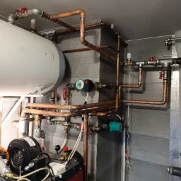 Złożona instalacja hydrauliczna z miedzianych rur, zaworów i pomp obiegowych, zamontowana w kotłowni z szarą, płytkowaną ścianą i bojlerem. Widoczne spawy i elementy kotła.