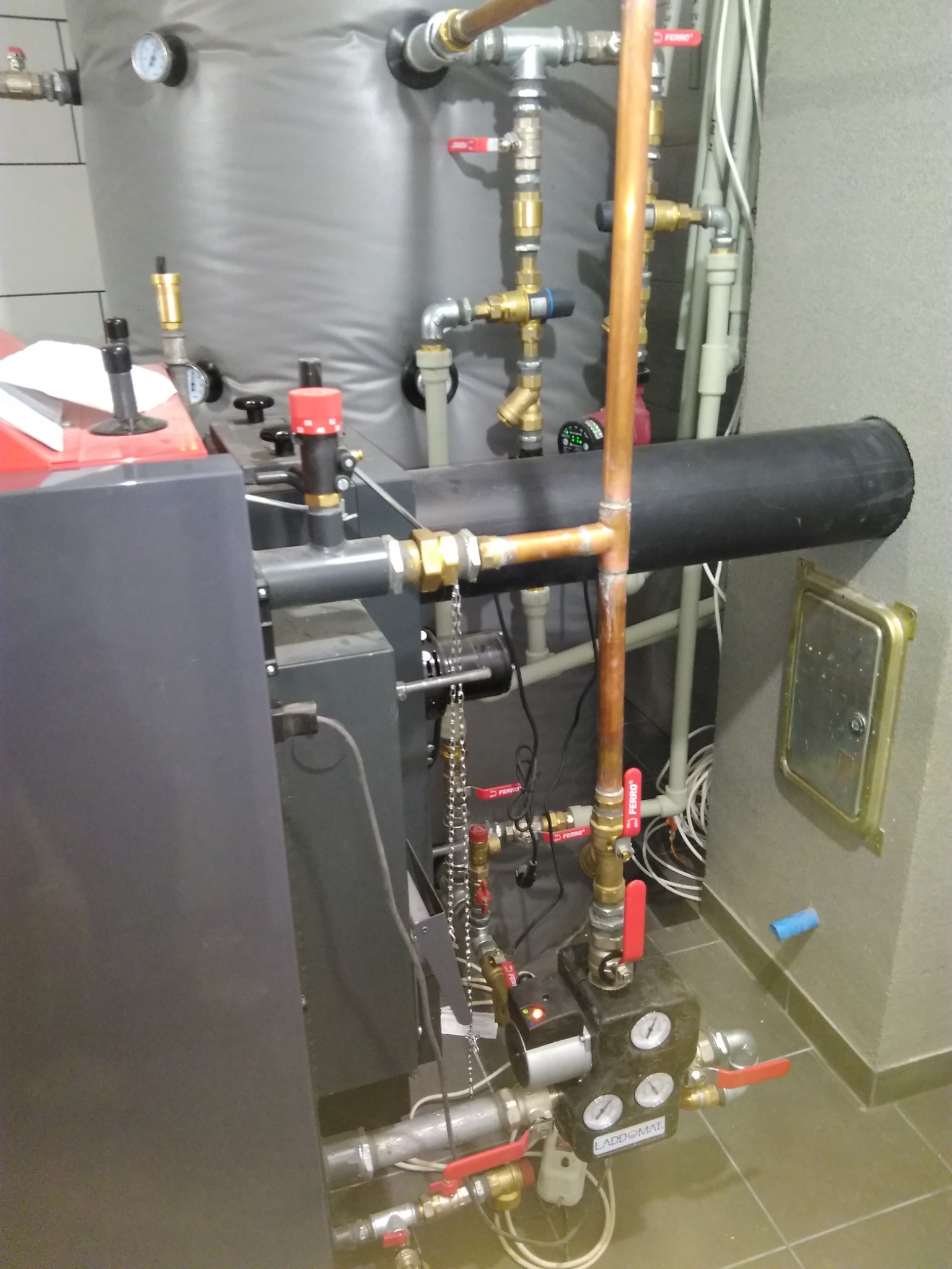 Szczegółowe ujęcie instalacji grzewczej z kotłem, rurami miedzianymi i zaworami Ferro. Widoczny czarny zbiornik izolowany i elementy pomiarowe LADDOMAT.