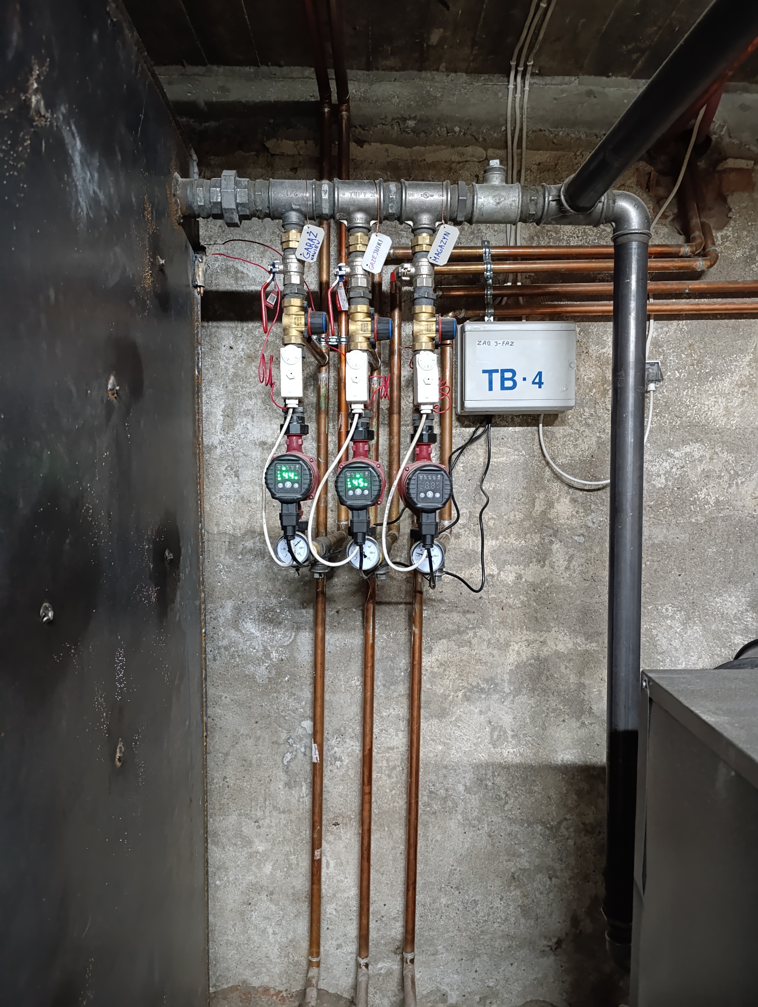 Instalacja hydrauliczna z trzema pompami obiegowymi z etykietami (garaż, grzejniki, magazyn) i rurami miedzianymi na szarej, surowej ścianie.