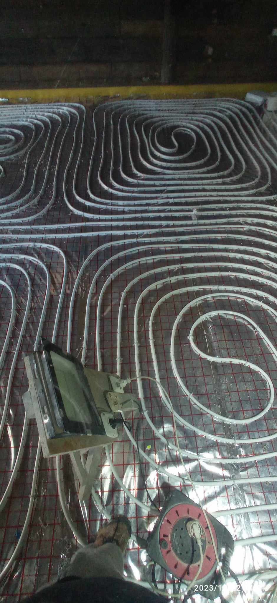 Instalacja ogrzewania podłogowego, rozłożone białe rury na folii z siatką, widoczne narzędzia pracy w Gródku. Ujęcie z góry, etap prac instalacyjnych.