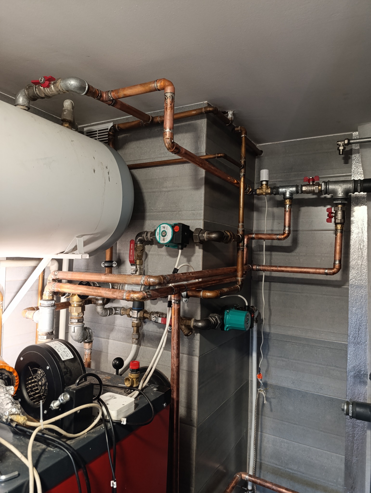 Złożona instalacja hydrauliczna z miedzianych rur, zaworów i pomp obiegowych, zamontowana w kotłowni z szarą, płytkowaną ścianą i bojlerem. Widoczne spawy i elementy kotła.