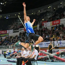 Skok o tyczce na zawodach lekkoatletycznych w hali, zawodnik w locie nad poprzeczką, w tle widownia i tablica wyników.