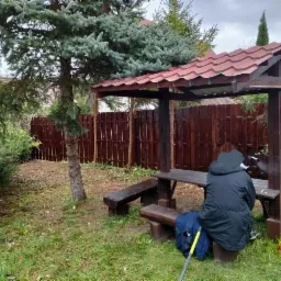 Altana ogrodowa z drewnianym ogrodzeniem w tle, osoba siedząca na ławce, butelka z napojem obok.