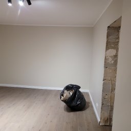 Ter-Bud - Wnętrze pokoju w trakcie remontu: świeżo pomalowane ściany w odcieniach beżu, jasna podłoga, widoczny fragment ściany po usunięciu futryny i czarny worek z odpadami budowlanymi w rogu.