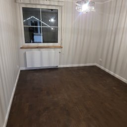 Ter-Bud - Pusty pokój po remoncie z ciemną drewnianą podłogą, białymi ścianami, grzejnikiem pod oknem i nowoczesną lampą wiszącą.