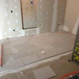 Ter-Bud - Układanie beżowych płytek podłogowych w trakcie prac wykończeniowych w łazience, widoczne poziomica, listwa i narzędzia glazurnicze.