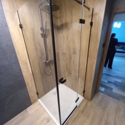 Ter-Bud - Kabina prysznicowa z czarnymi profilami i drewnopodobnymi płytkami na ścianach, widok z boku, z fragmentem osoby w tle.