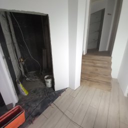 Ter-Bud - Wnętrze w trakcie układania podłogi z płytek imitujących drewno, widoczne narzędzia glazurnicze, wiadro z klejem i niezagospodarowane wejście do łazienki z widoczną instalacją elektryczną.